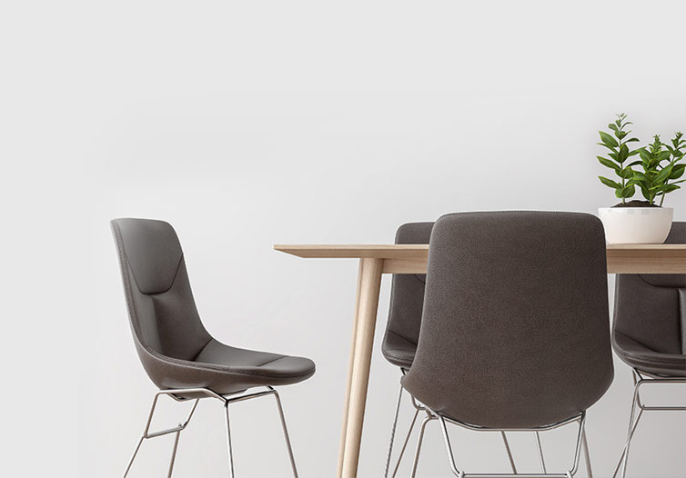 A modern dining area featuring a wooden table with sleek legs and three black, cushioned chairs with metal frames. A white planter with a green plant decorates the table. The background is a plain light gray wall.