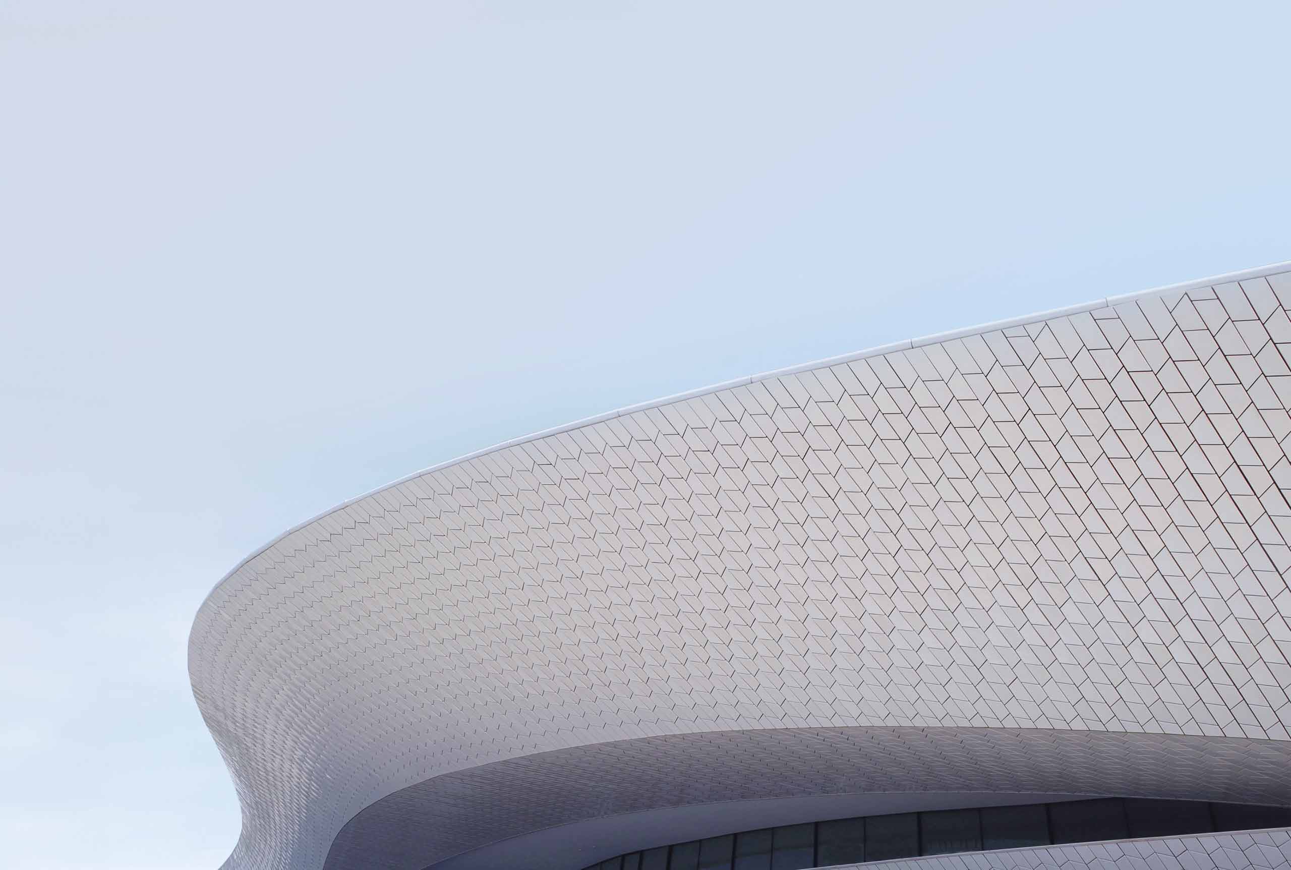 Curved modern building with a textured facade against a clear blue sky. The surface features a grid pattern, creating a sleek, futuristic appearance.