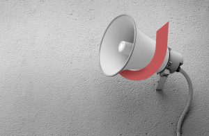 A gray wall-mounted loudspeaker with a red, curved shape encircling it. The speaker is on the right side of the image