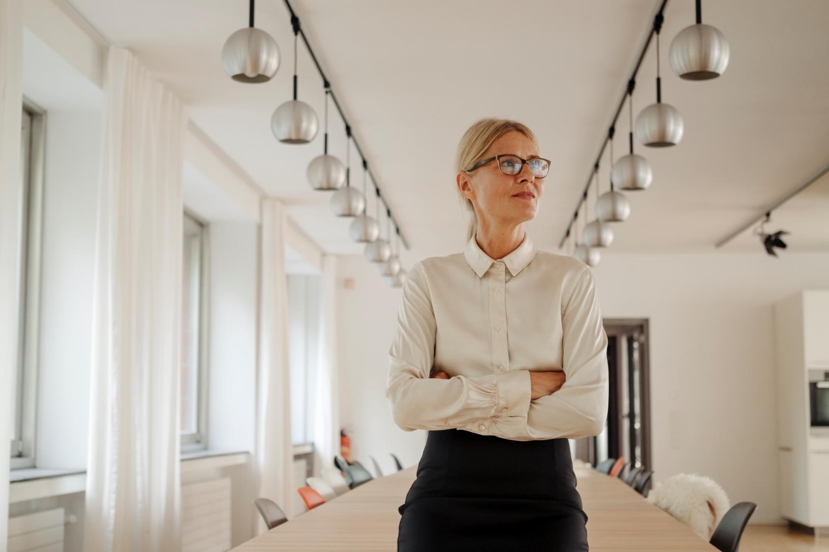 (direct upload) A woman in professional attire stands confidently with arms crossed in a modern conference room. The room has a long table, multiple hanging lights, and large windows with curtains, allowing natural light in.