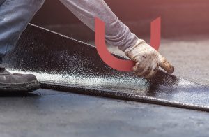 A worker in gloves is applying a black waterproof sealant to a roof surface.