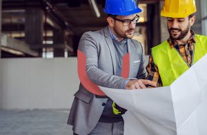 Two men in hard hats review blueprints at a construction site. One wears a gray suit and blue helmet, while the other wears a yellow helmet and high-visibility vest. They are in a partially completed building.