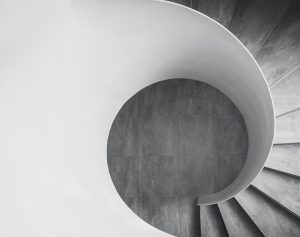 A black and white image of a spiral staircase viewed from above, showcasing a curving design. The center reveals a tiled floor, and the stairs have a smooth, modern finish. The composition emphasizes the geometric lines and shadows.