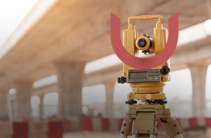 A theodolite set up in front of a large, blurred concrete bridge under construction. The equipment is yellow with electronic controls, focusing on precise measurements. The scene suggests a surveying project.