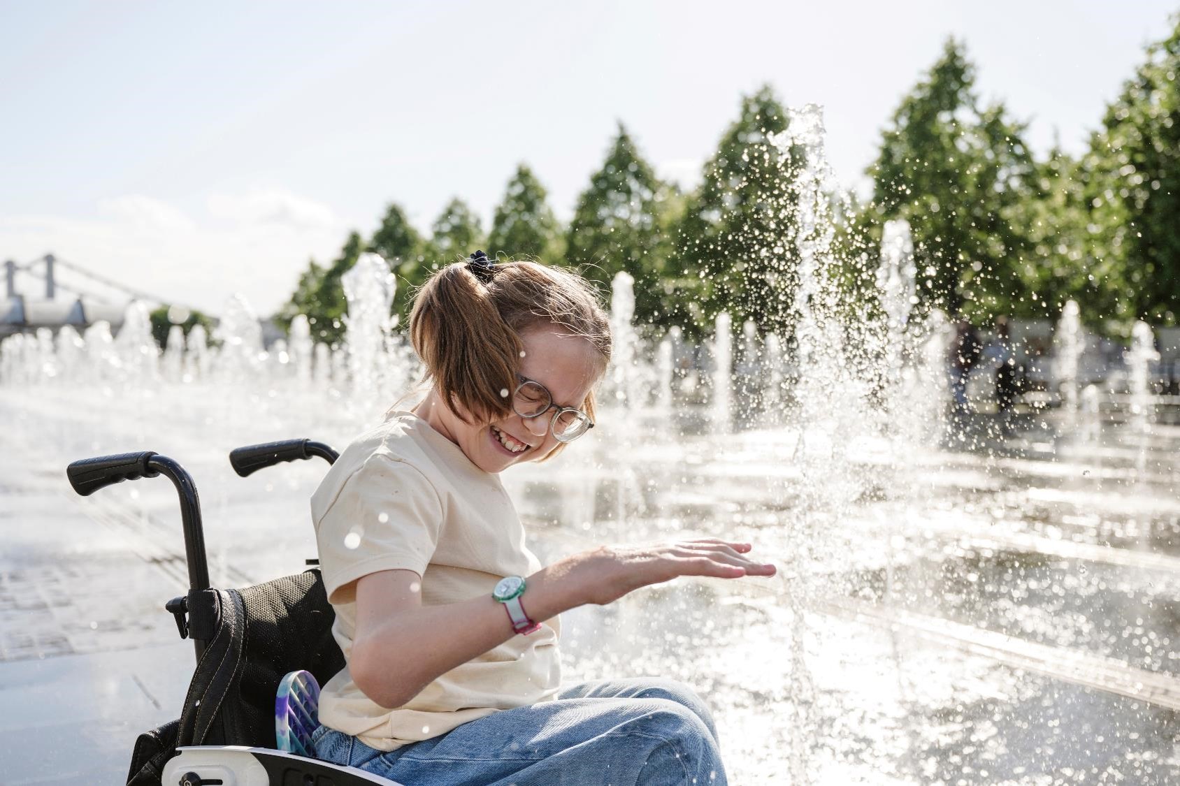 A young girl in a wheelchair smiles and reaches out to touch the water from a fountain on a sunny day. She is wearing glasses and a casual outfit. The background features trees and a clear sky.