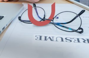 A pair of eyeglasses rests on an upside-down document labeled Resume. Nearby are a laptop and a smartphone on a light wood surface. A red, curved shape overlays the image.