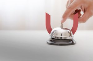 A hand pressing a silver service bell on a white surface. The setting appears minimalist with soft lighting.