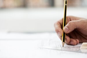 A close up of an architect's hand drawing a line on an architectural drawing utilizing a plastic ruler.