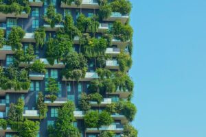 A modern high-rise building with numerous balconies, each adorned with lush green plants and trees, set against a clear blue sky.