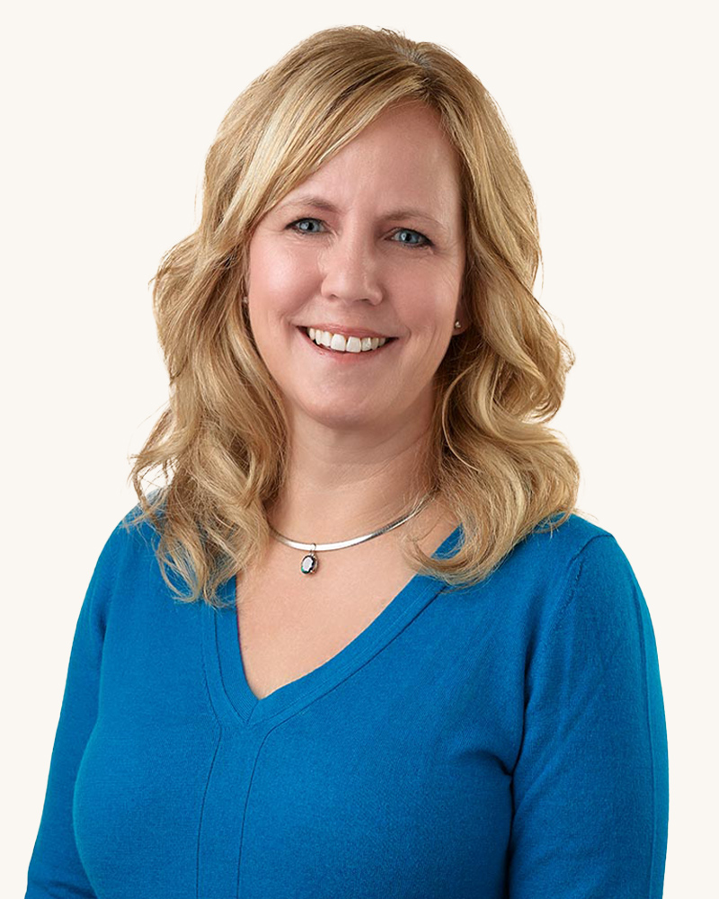 A woman with blonde wavy hair smiles, wearing a blue V-neck top and a silver necklace with a pendant. The background is plain white.