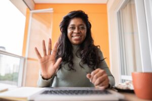 A woman with long dark hair sits at a desk in front of a laptop, smiling and waving at the camera. She is indoors with an orange wall and a window behind her. A cup and a small plate are on the desk next to her.