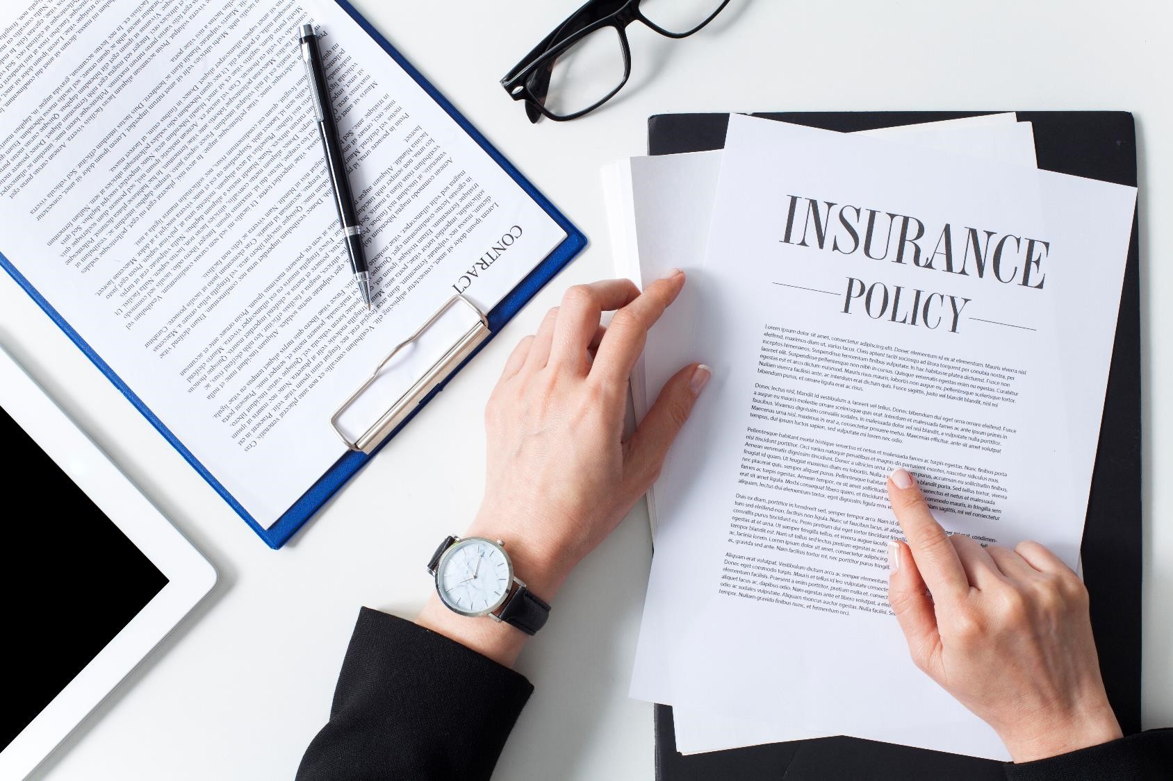 A person is reviewing an insurance policy document on a desk. Nearby are a contract, a pen, a pair of glasses, a tablet, and a wristwatch. The setting is a professional work environment with emphasis on paperwork and decision-making.