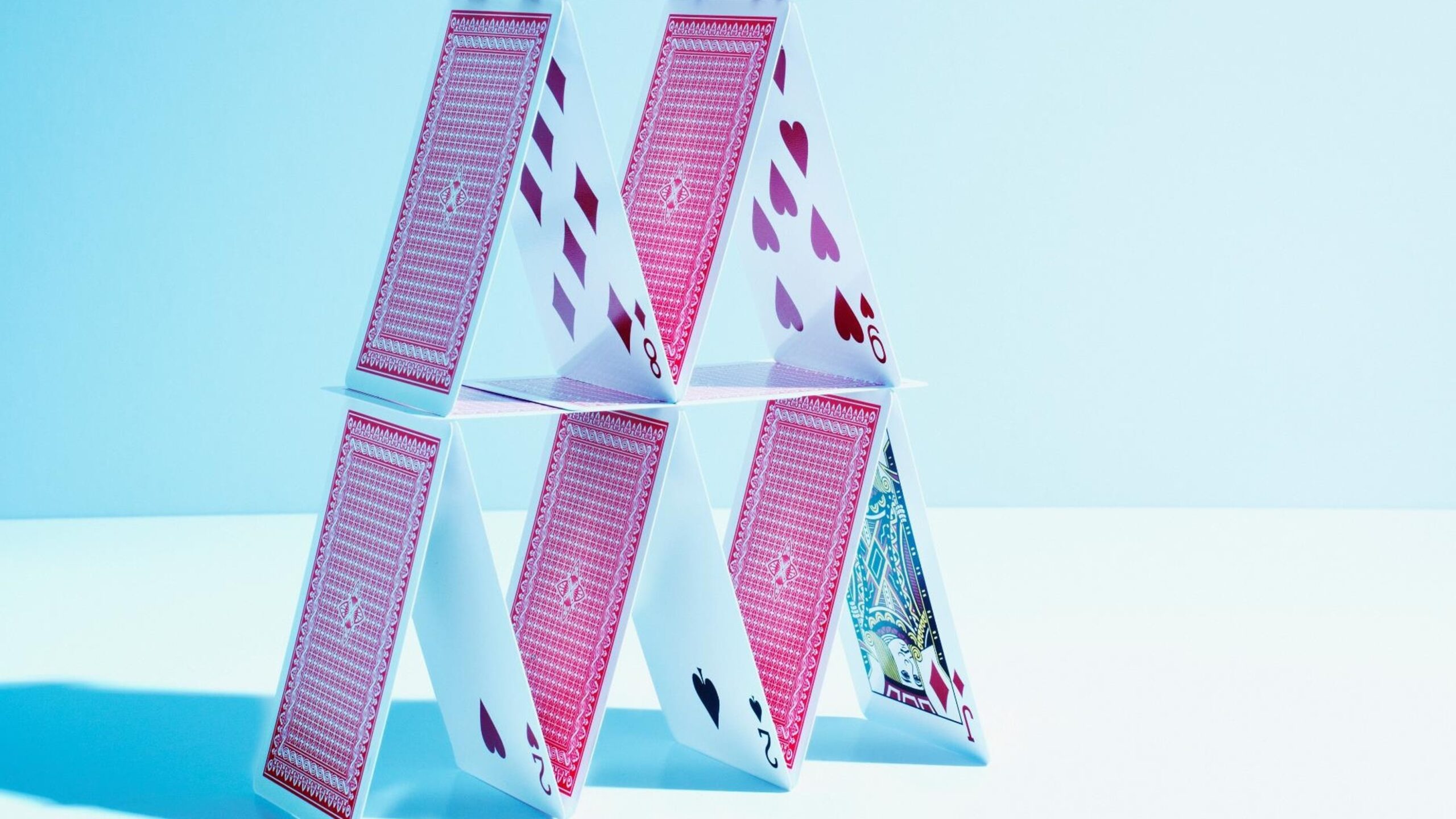 A house of cards with two levels, built using standard playing cards with red backs and visible suits like hearts, spades, and clubs. The structure is set against a light blue background, casting soft shadows.