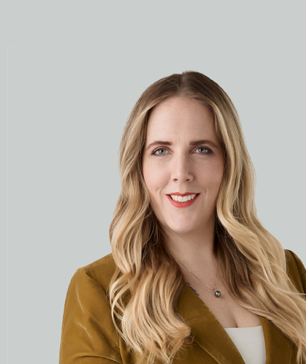 A woman with long, blonde hair smiles at the camera. She is wearing a brown jacket over a white top, and her background is a solid light gray.