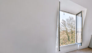 A minimalist white room with a large open window, revealing a view of bare trees and a clear sky outside. The window tilts inward, allowing natural light to fill the space.