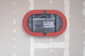 A hole in drywall reveals a network of cables and wiring. The central cutout is framed by a red oval, drawing attention to the exposed electrical setup against a background of unfinished wall.